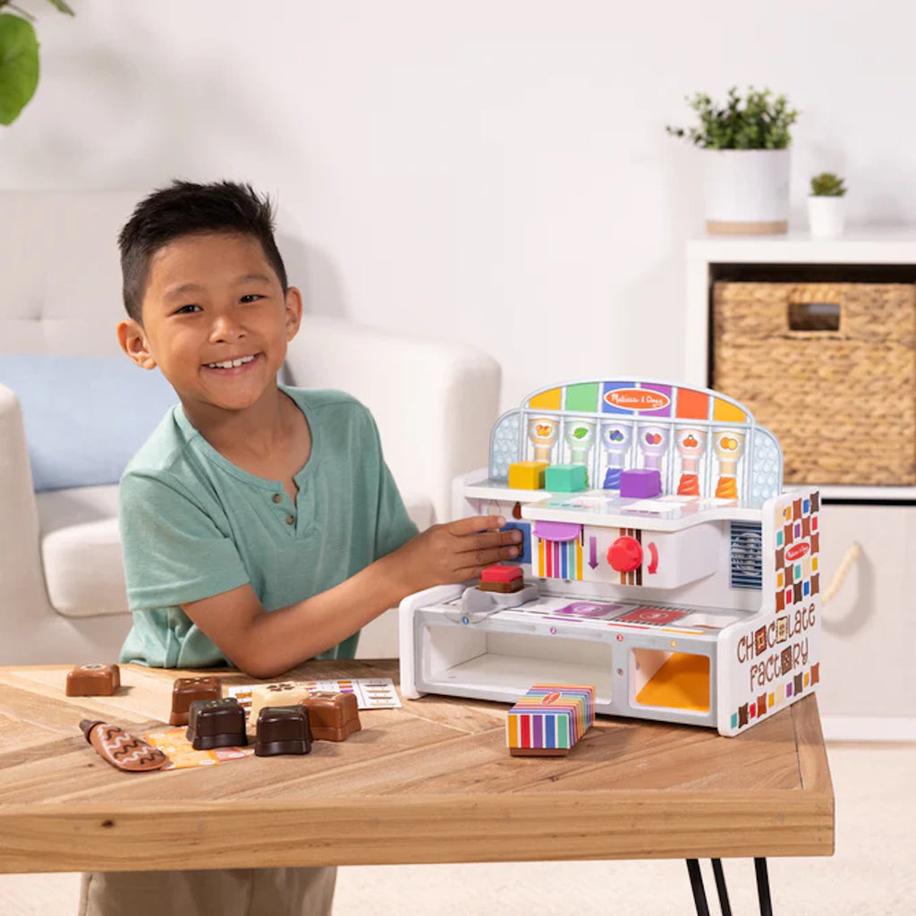 Child playing with a toy Chocolate Factory set on a table, smiling at the camera.